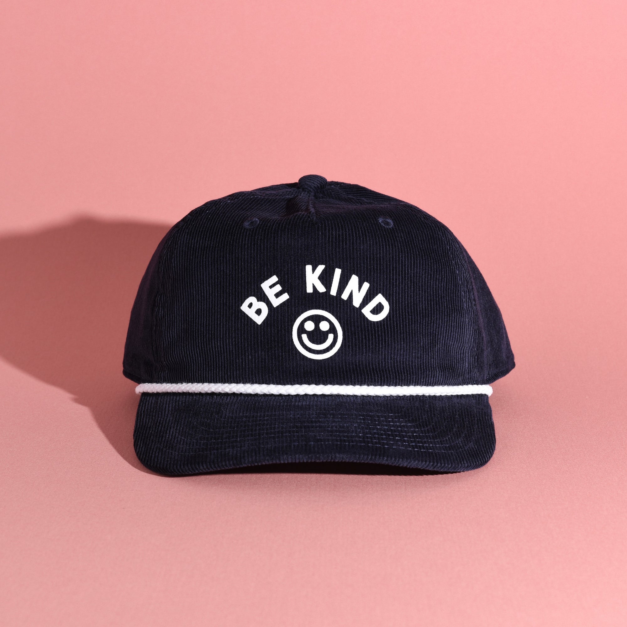 Navy blue corduroy cap with 'BE KIND' text and smiley face on a pink background