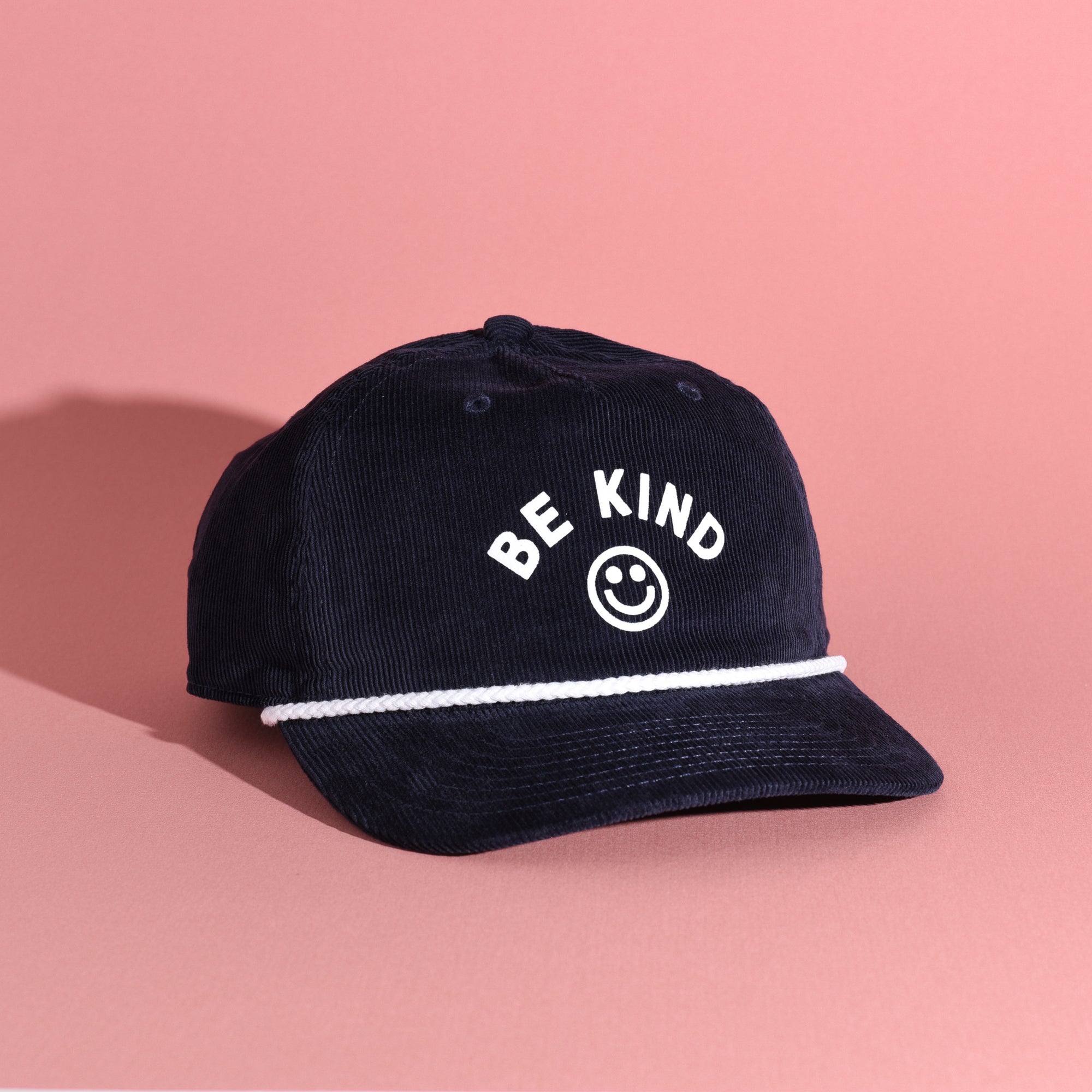 Navy blue corduroy cap with 'BE KIND' text and smiley face on a pink background