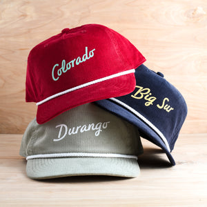 Three baseball caps with 'Colorado', 'Big Sur', and 'Durango' text on a wooden surface. These are customizable hats.